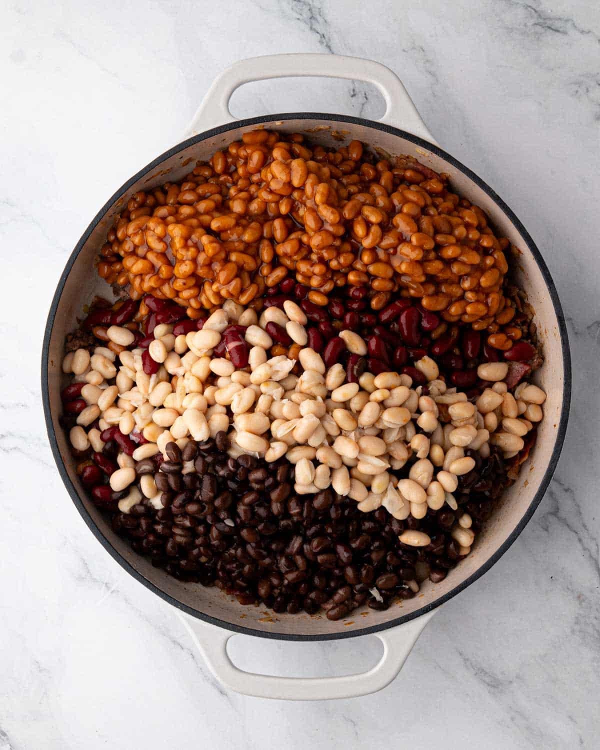 A white braiser with four types of beans added: baked beans, kidney beans, pinto beans, and black beans.