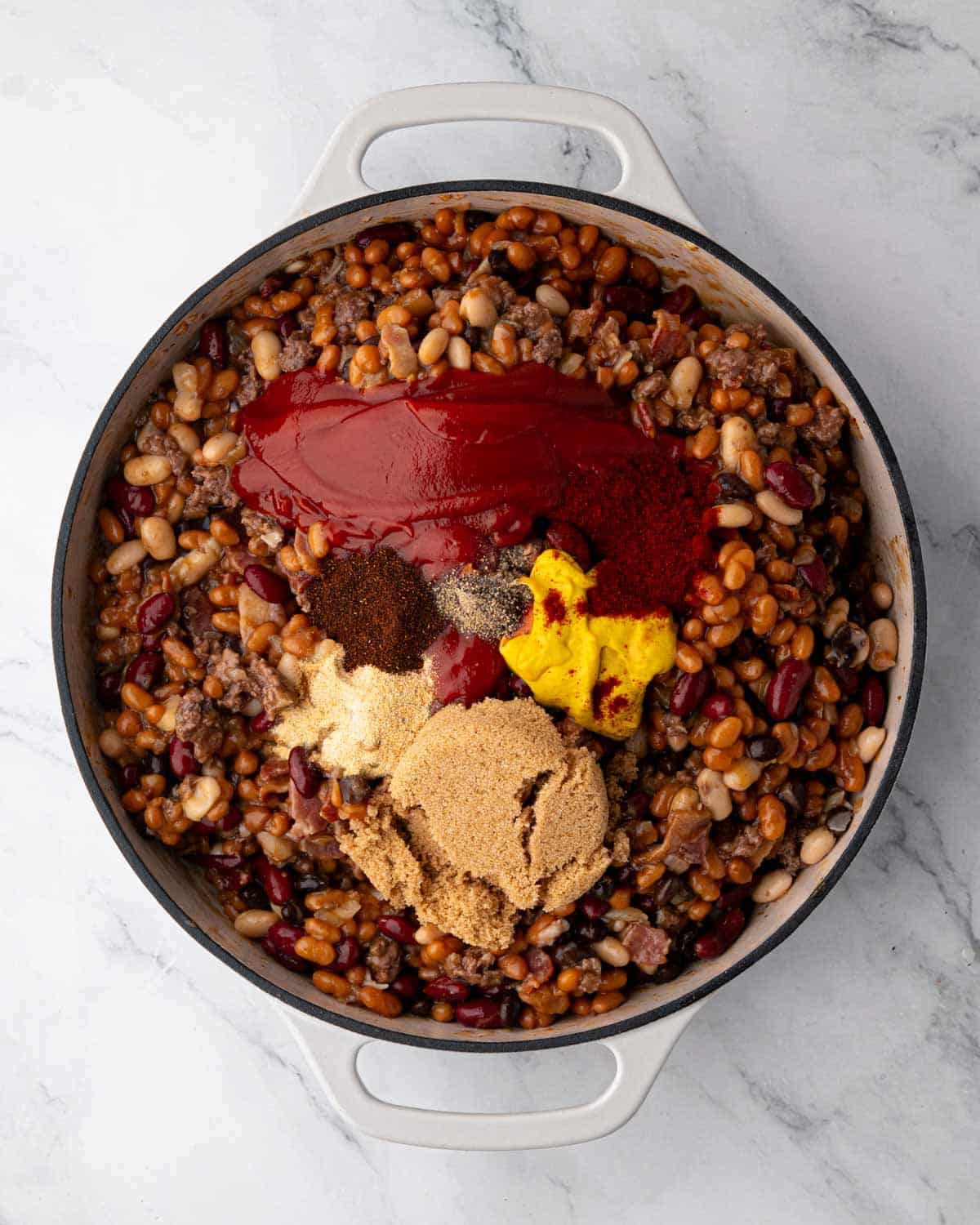 A white braiser with bean mixture and ketchup, mustard, brown sugar, and spices on top.