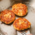 A skillet with fried mashed potato patties with a hand using a spatula to put underneath one patty.