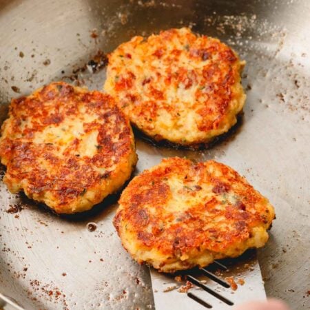 A skillet with fried mashed potato patties with a hand using a spatula to put underneath one patty.