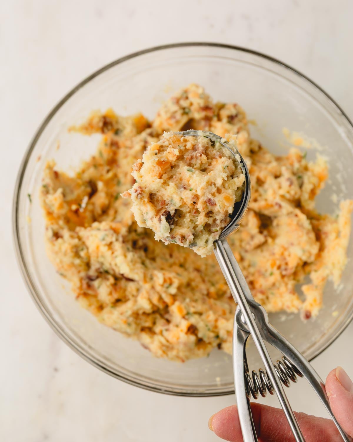A glass bowl with fried mashed potato patty mixture and a stainless steel scoop with some of the mixture in it.