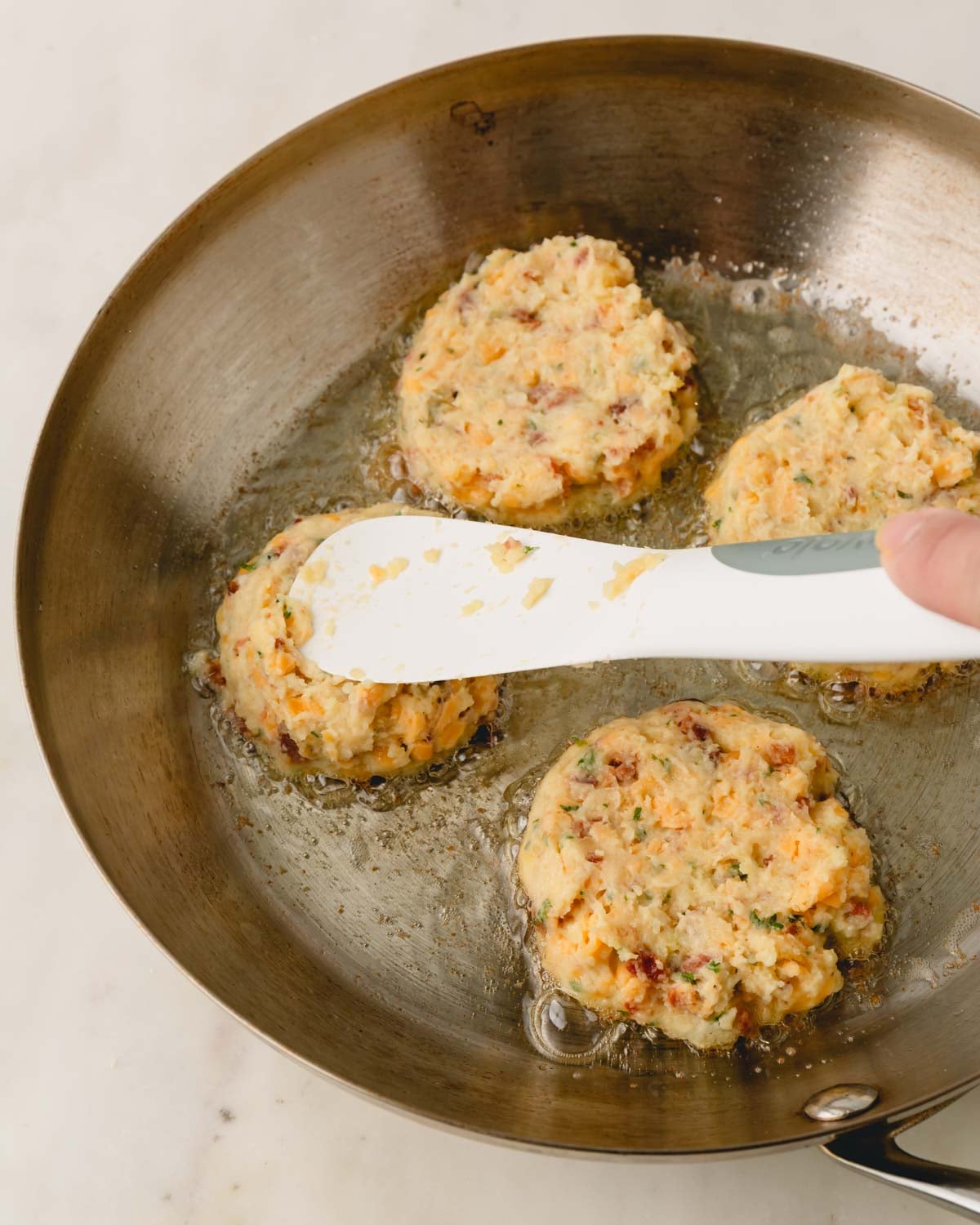 A skillet cooking mashed potato patties with a hand pressing down the patties with a white spatula.