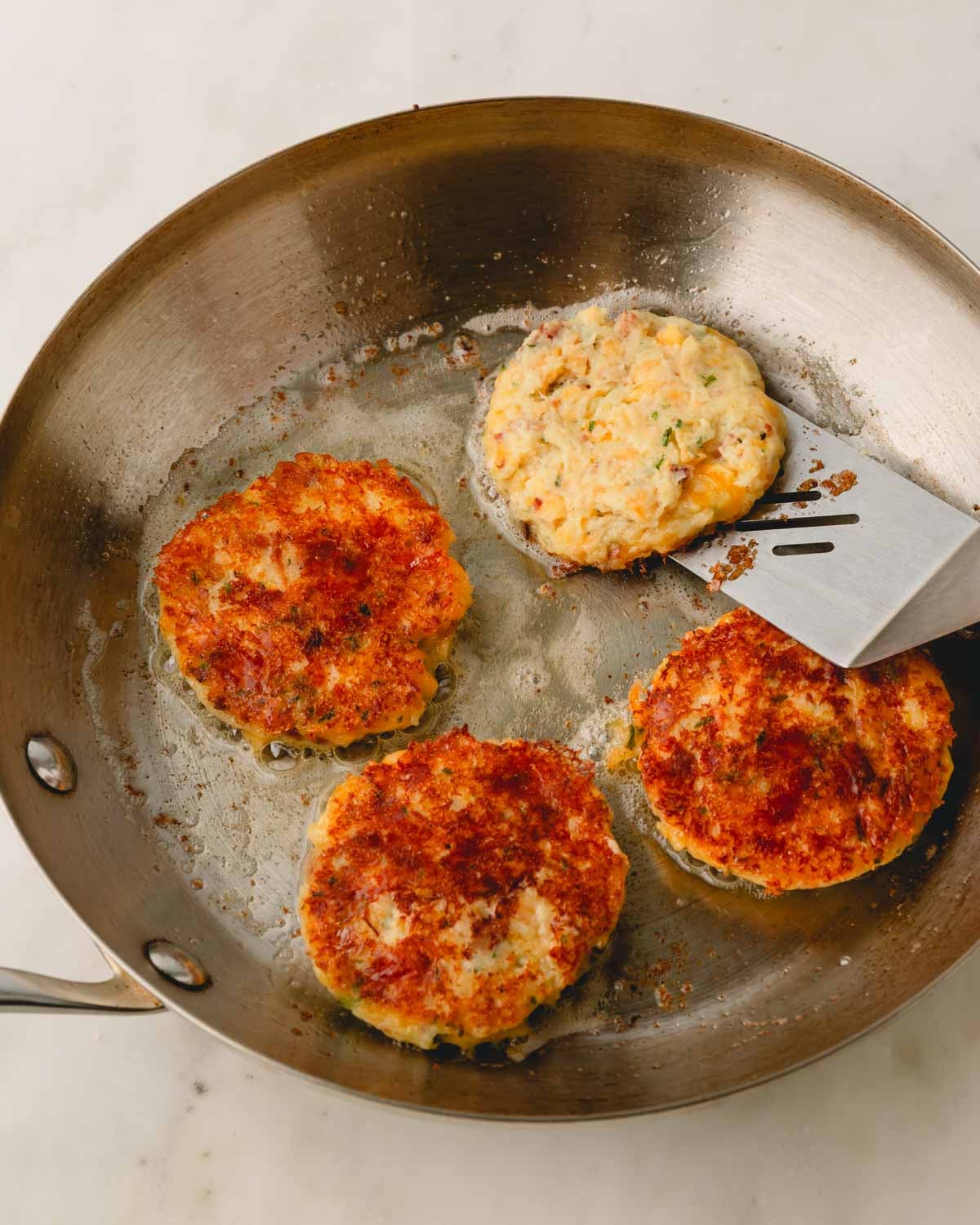 A skillet with mashed potato patties, three are flipped and one has a metal spatula underneath about to be flipped.