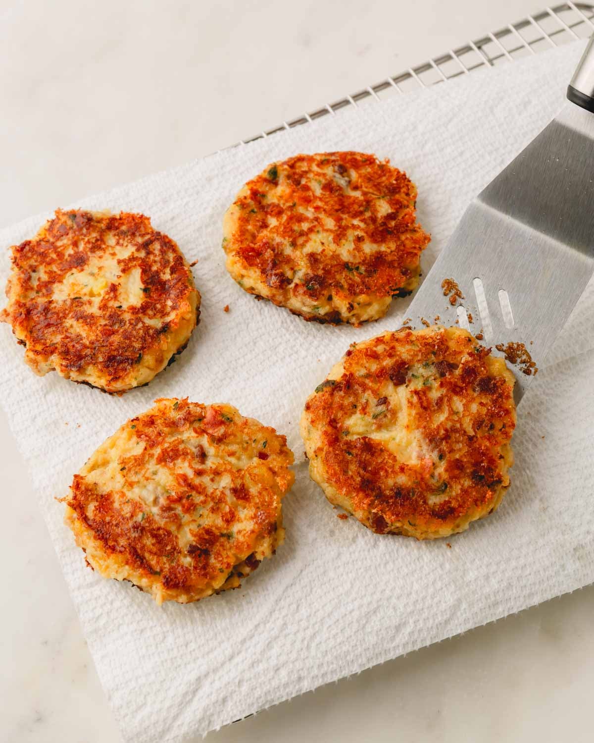 Mashed potato patties are draining on a paper towel over a wire rack. A metal spatula is lifting one patty up.
