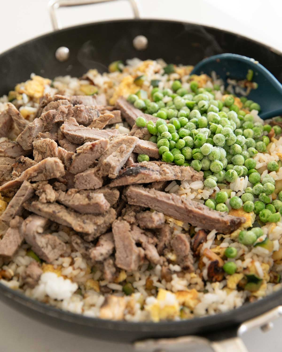 A skillet with steak, peas, and fried rice cooking with a blue spoon.