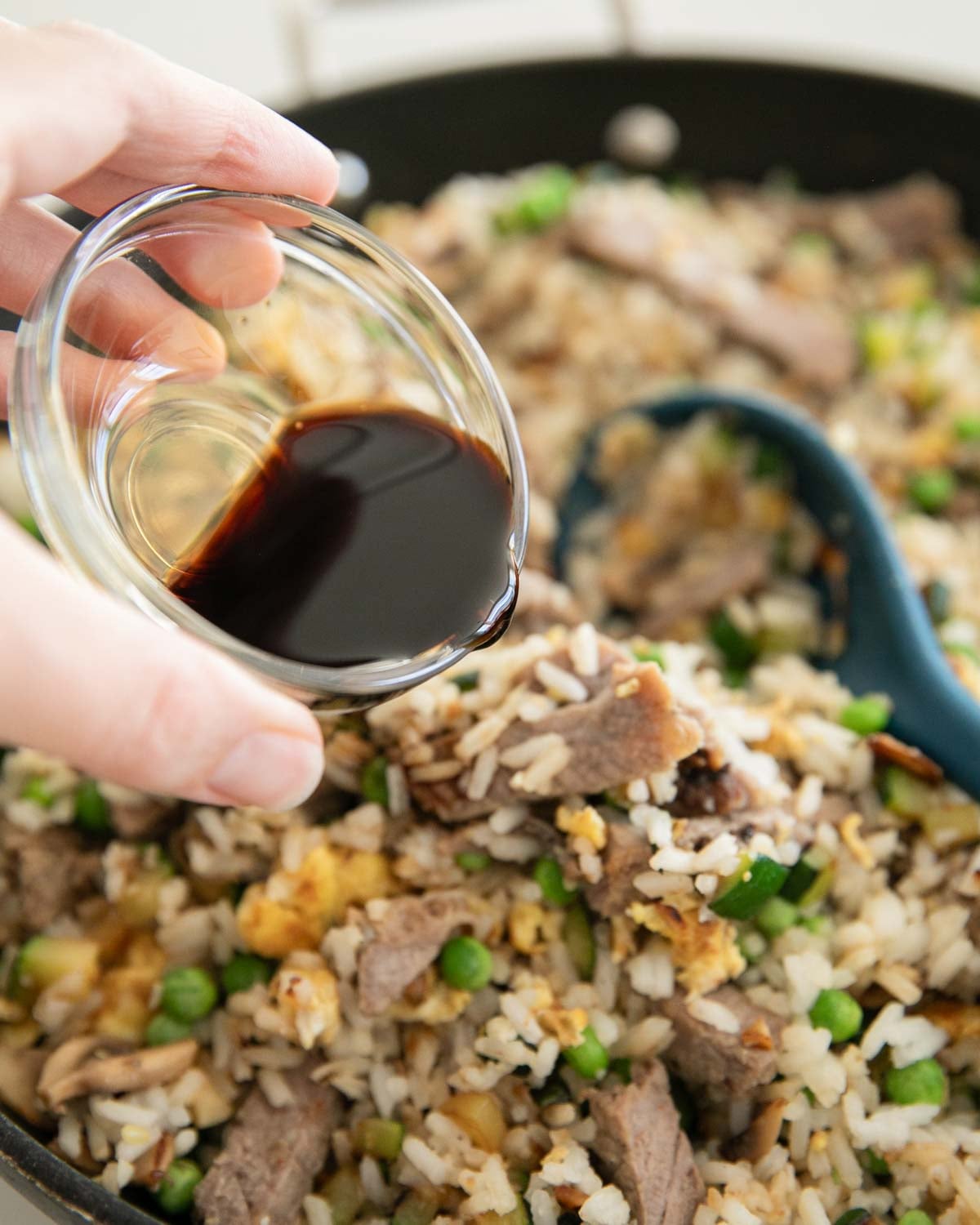 A hand pouring soy sauce into a skillet of steak fried rice in a glass bowl.