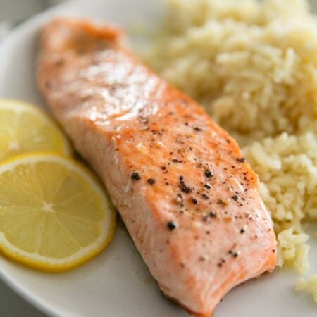 A piece of steelhead trout on a white plate with rice and lemon slices.