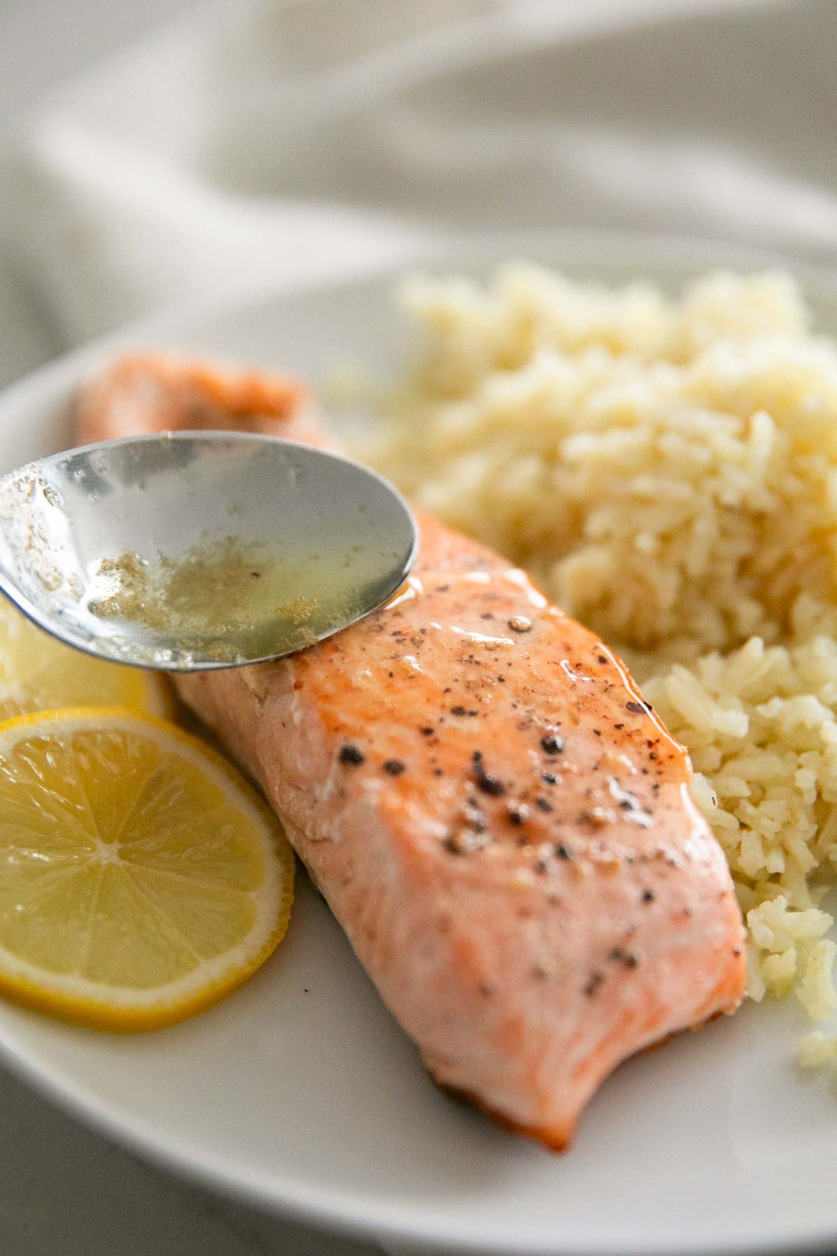 A spoon drizzling the lemon thyme sauce over the steelhead trout fillet with lemons slices and rice on a white plate.