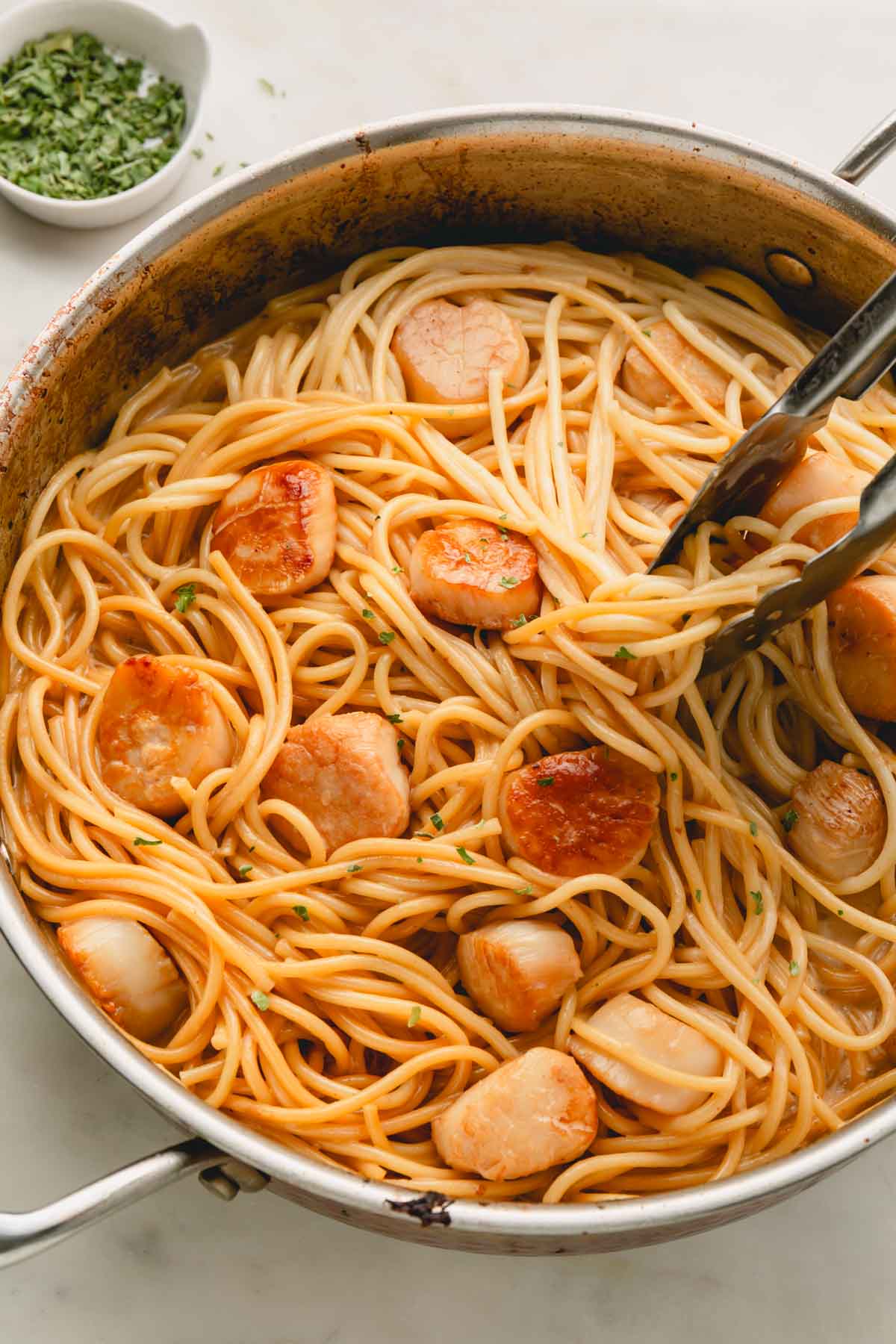 Tongs tossing scallop pasta in a large skillet.