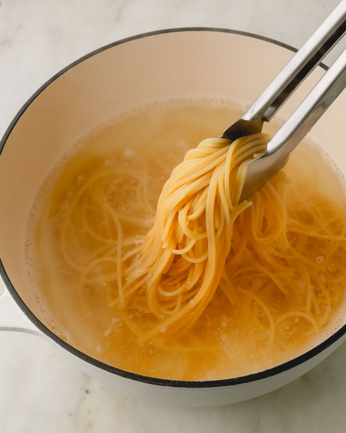 Tongs holding noodles being boiled in a white pot.