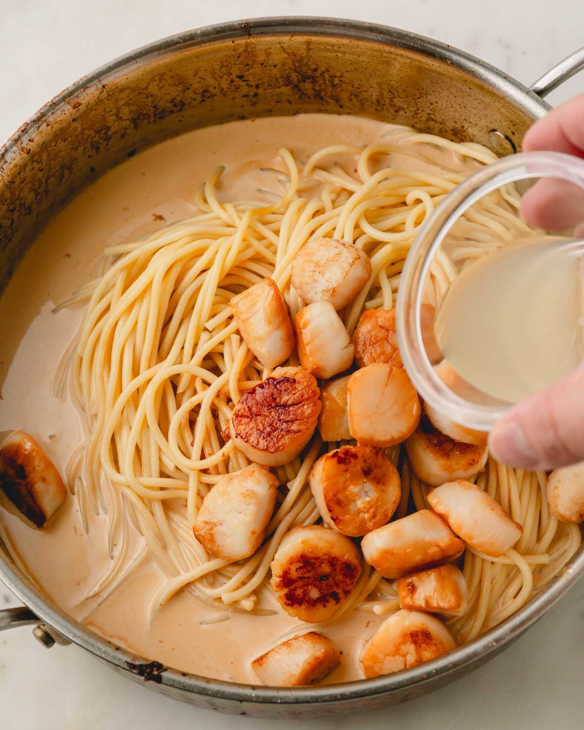 A hand holding a glass bowl of lemon juice being added to scallop pasta.