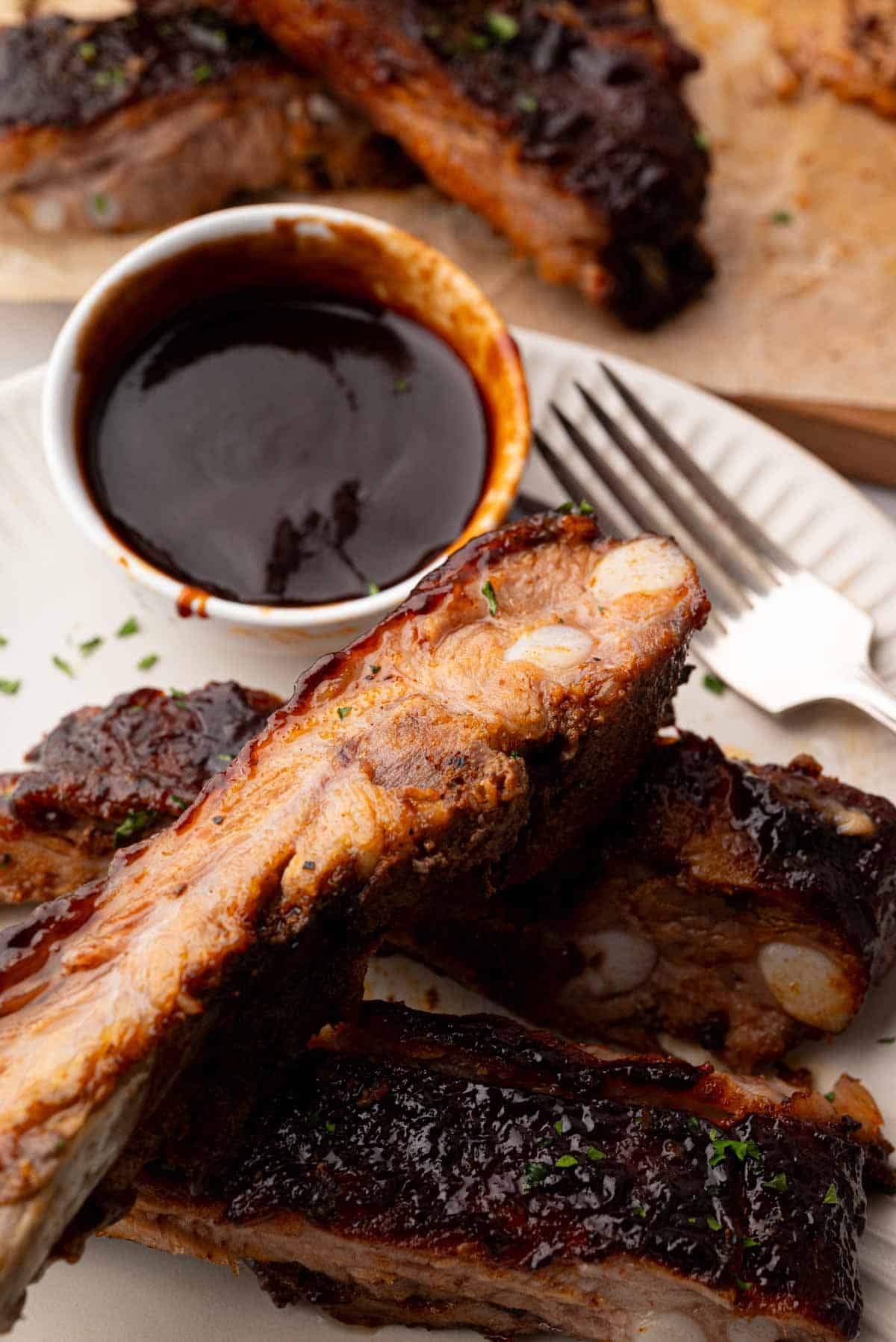 A close up image of a cut and stacked St. Louis spare ribs on a white plate with a white bowl of barbecue sauce and a fork on the side.
