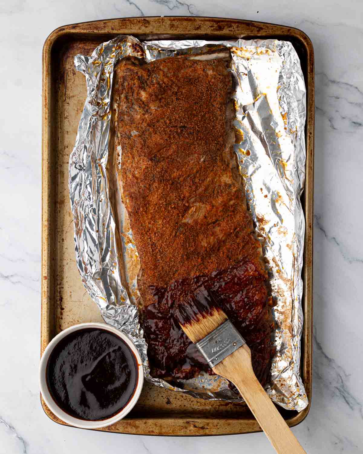 Baked St. Louis ribs being brushed with bbq sauce on a foil-lined baking sheet. The barbecue sauce is in a small white bowl.