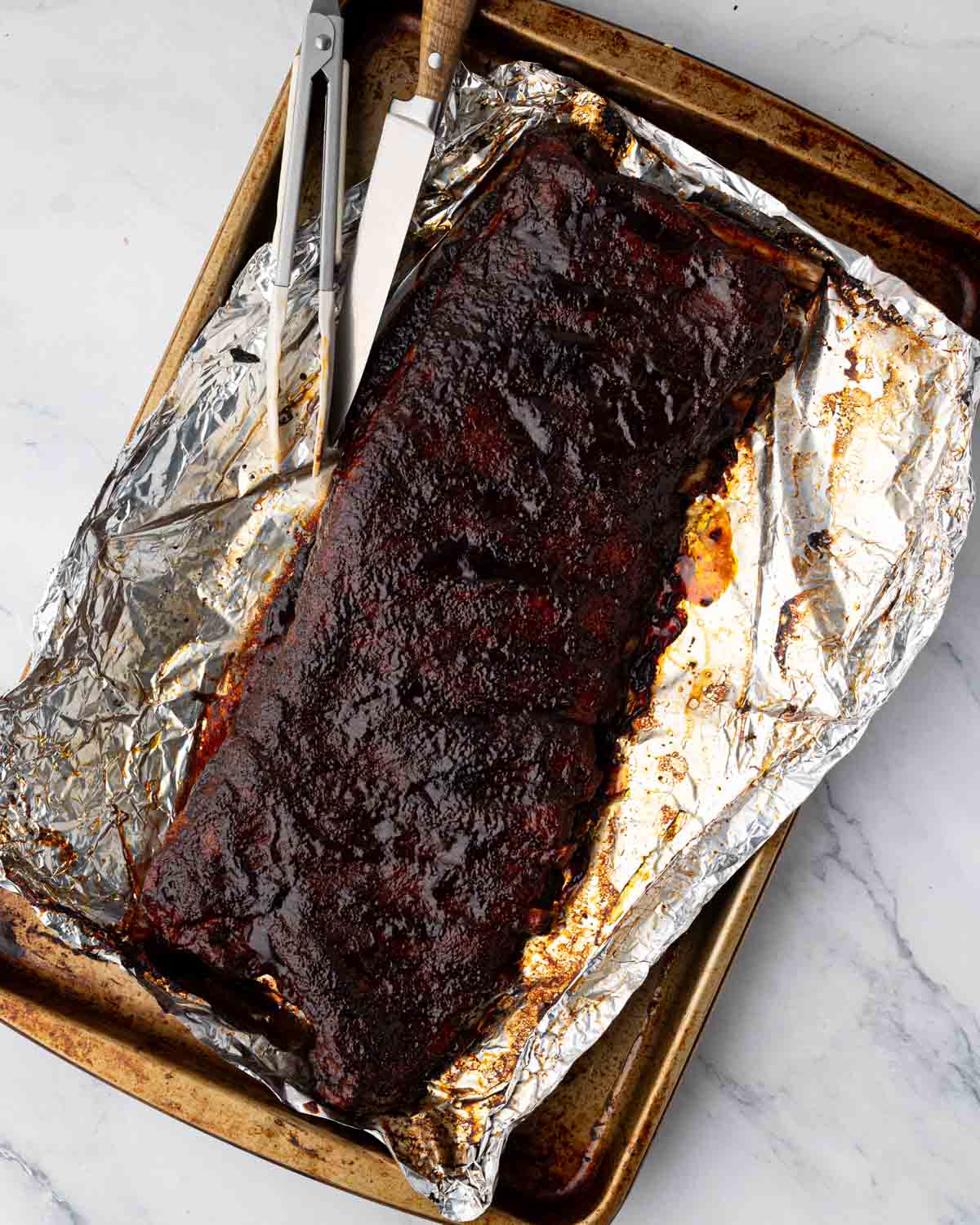 A foil-lined baking sheet with St. Louis ribs baked and sauced ready to cut. A knife and tongs are to the side.