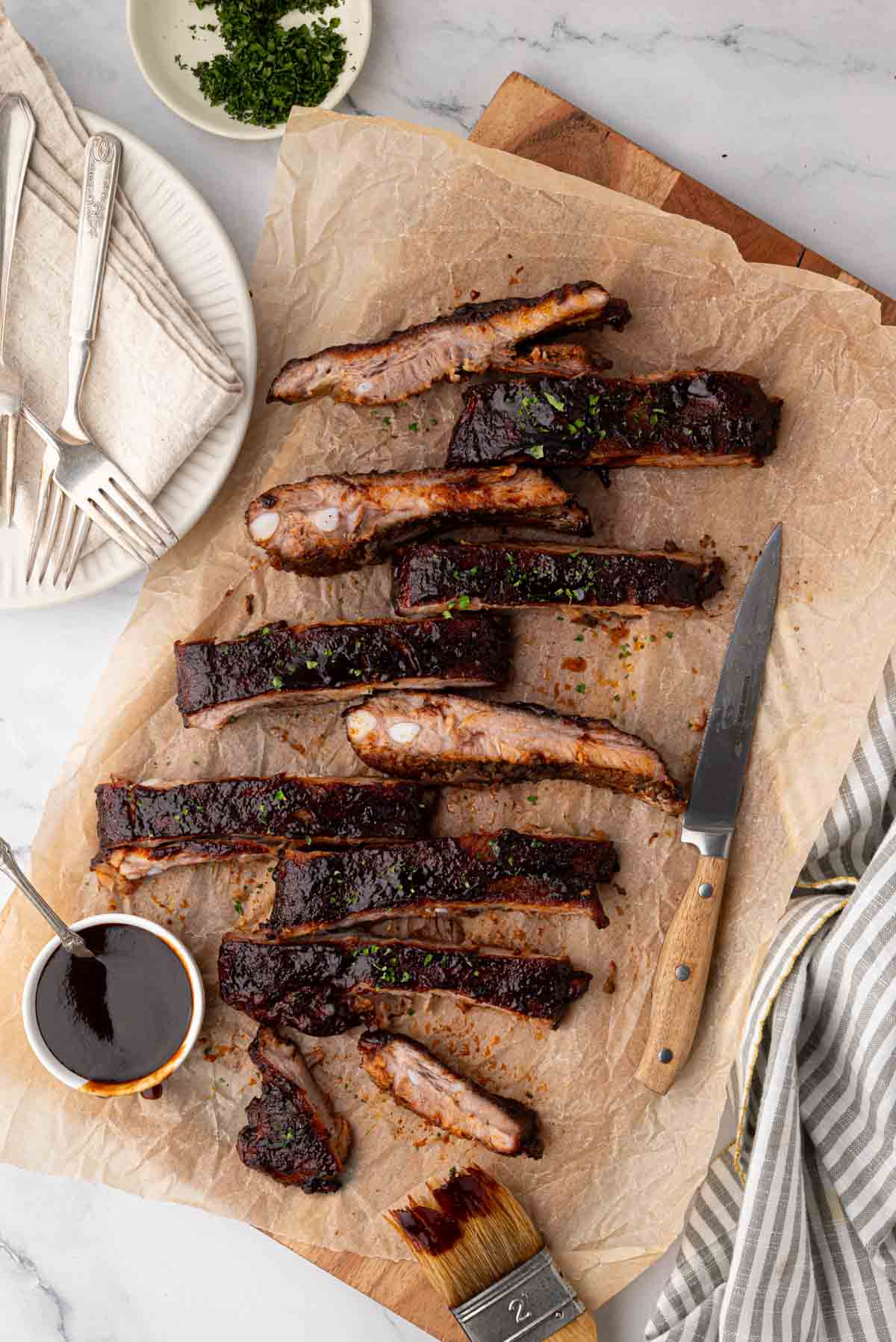 A cutting board with parchment paper and sliced St. Louis bbq ribs on a wooden cutting board. White plates, napkins, and silverware are to the left.