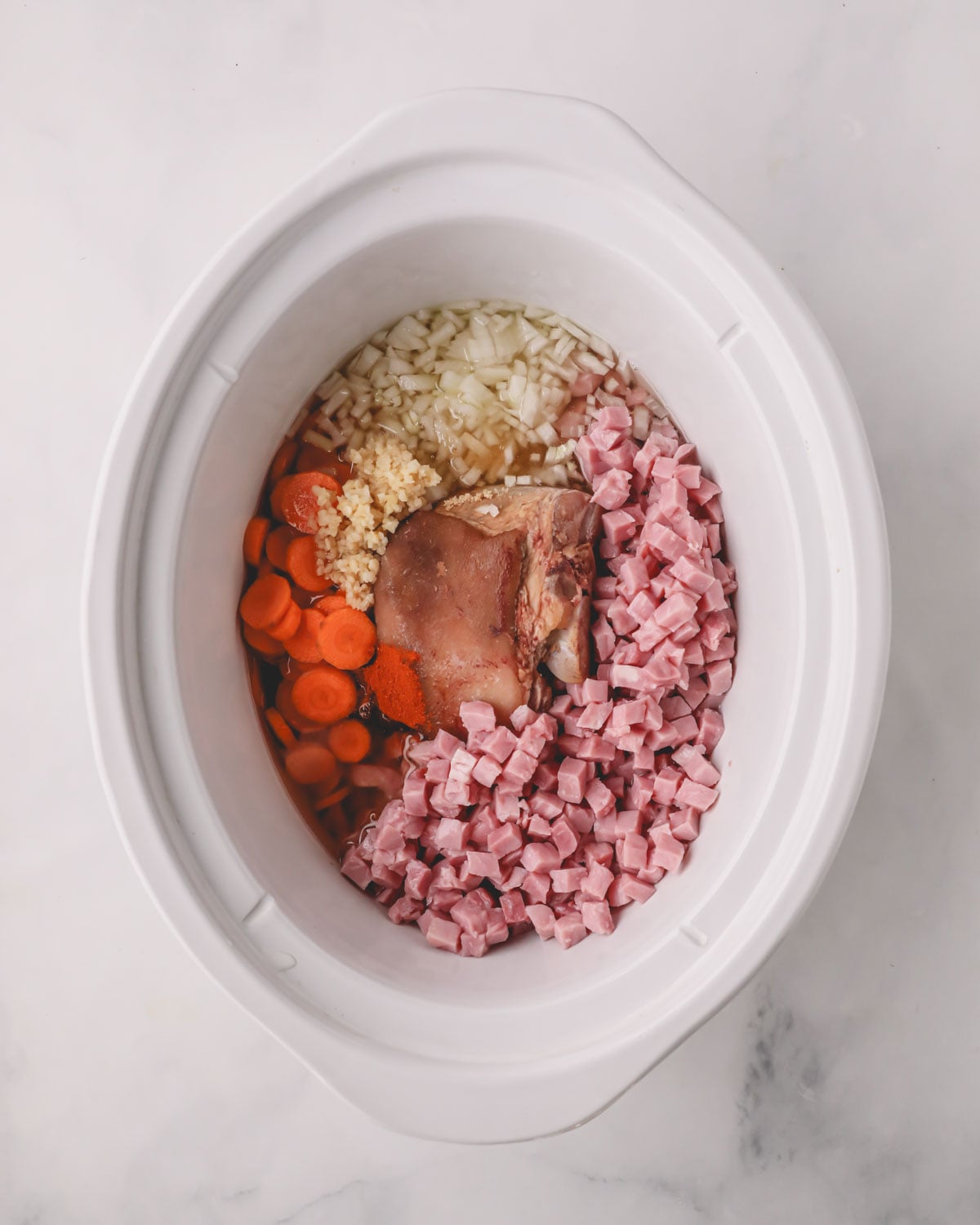 A white slow cooker with aromatic ingredients and a ham hock in the center.