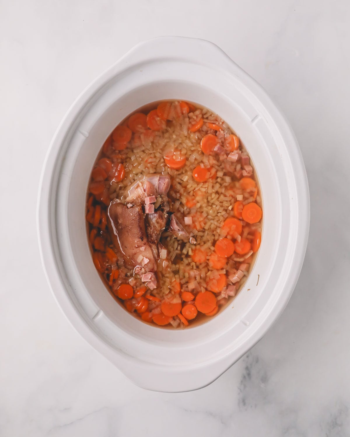 A white slow cooker with ham hock, vegetables, and chicken stock.