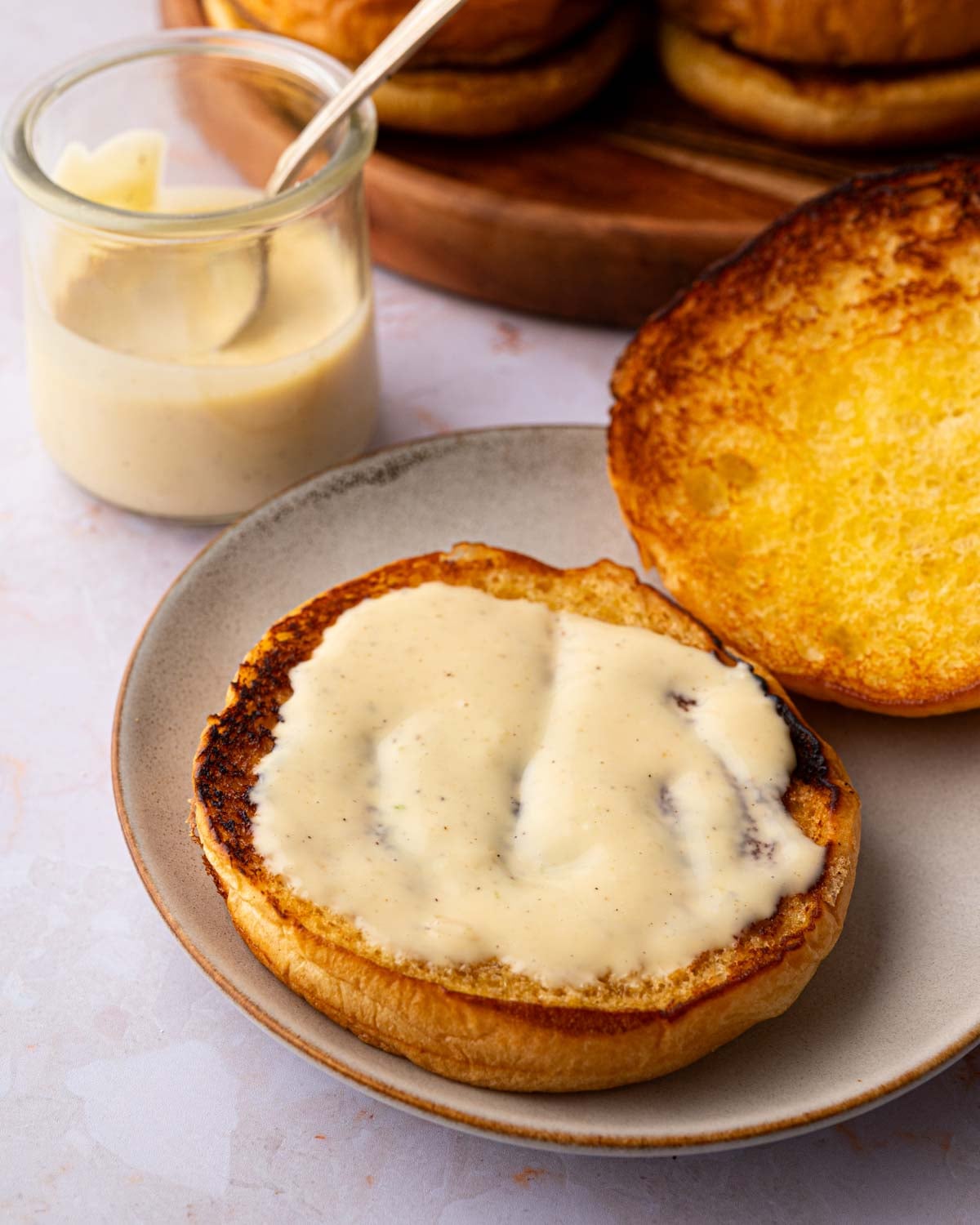 Toasted hamburger bun with honey mustard aioli on a gray plate and a glass jar of more aioli with a spoon in the background.