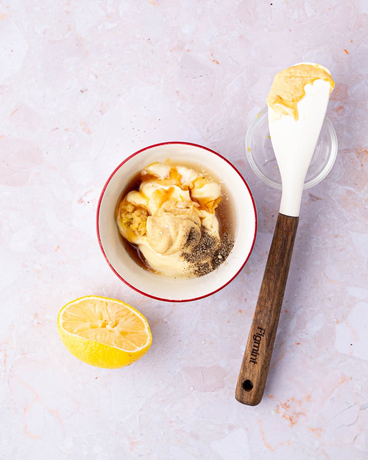 A white bowl with ingredients for honey mustard aioli. A white spatula is to the side with a glass bowl and a squeezed lemon half.
