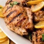 Close up of grilled lemon chicken on a white platter with lemon slices.