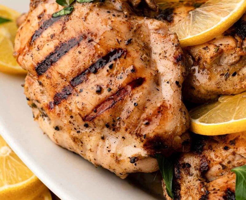 Close up of grilled lemon chicken on a white platter with lemon slices.