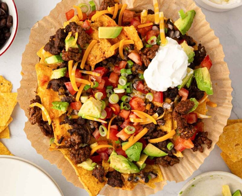 Overhead image of air fryer nachos in parchment paper on a plate with toppings around it.
