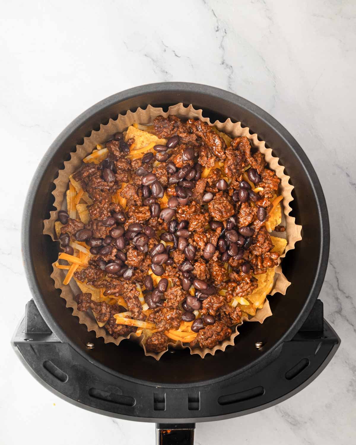 An air fryer basket with parchment liner, tortilla chips, ground beef, and black beans.