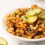A white plate with cheeseburger pasta with dill pickle slices on top.