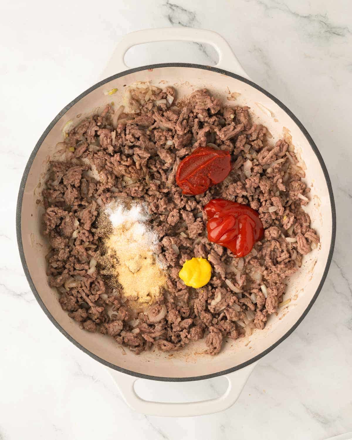 A white skillet with cooked ground beef, spices, ketchup, tomato paste, and mustard.