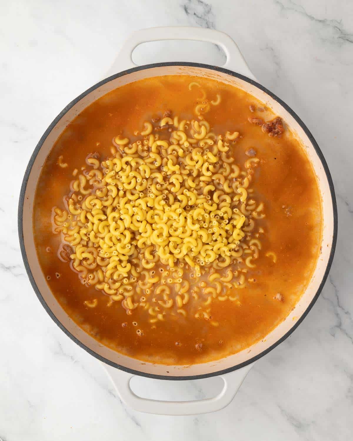 A white skillet with browned beef, spices, broth, and macaroni noodles.