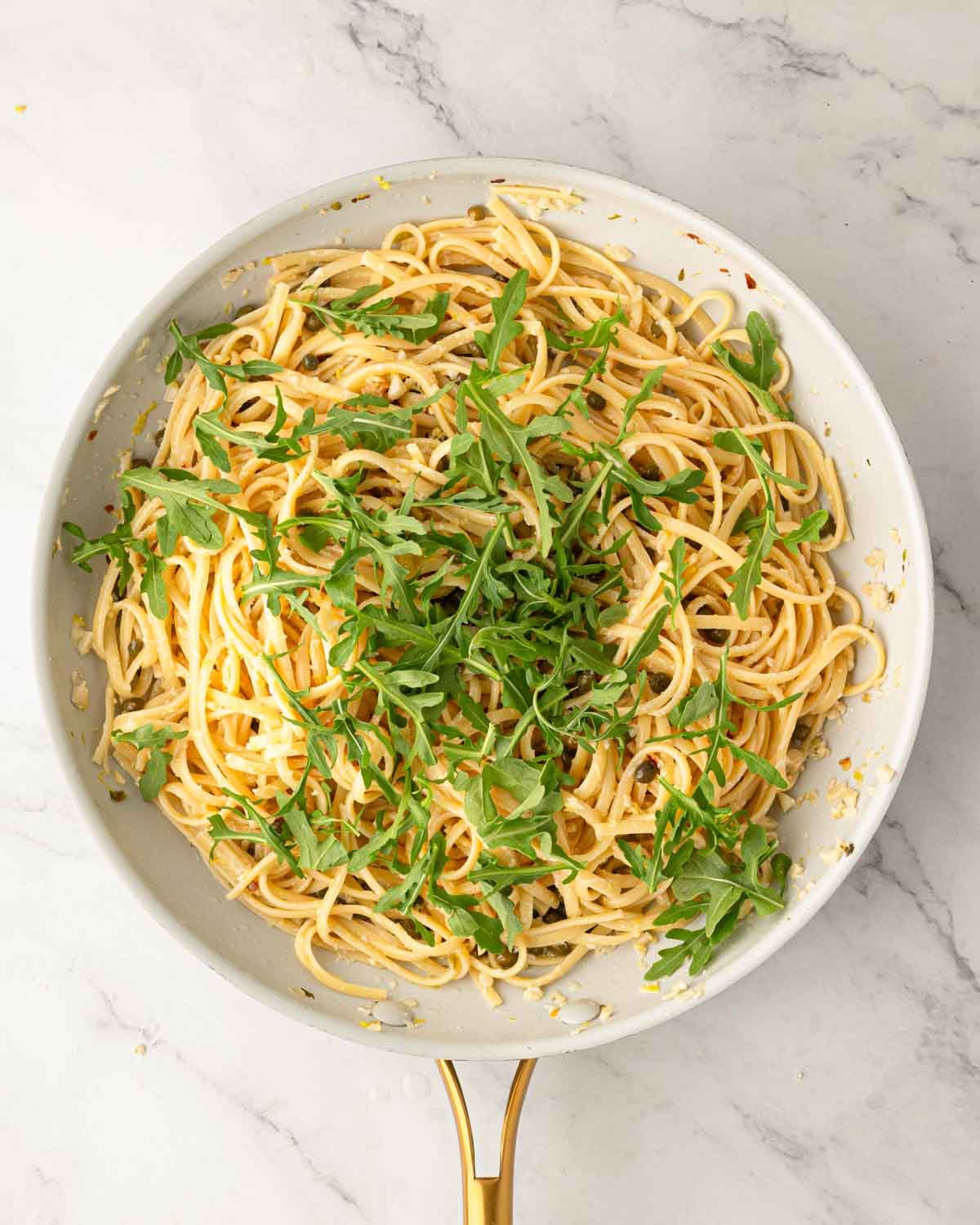 A white skillet with lemon caper pasta and arugula on top.