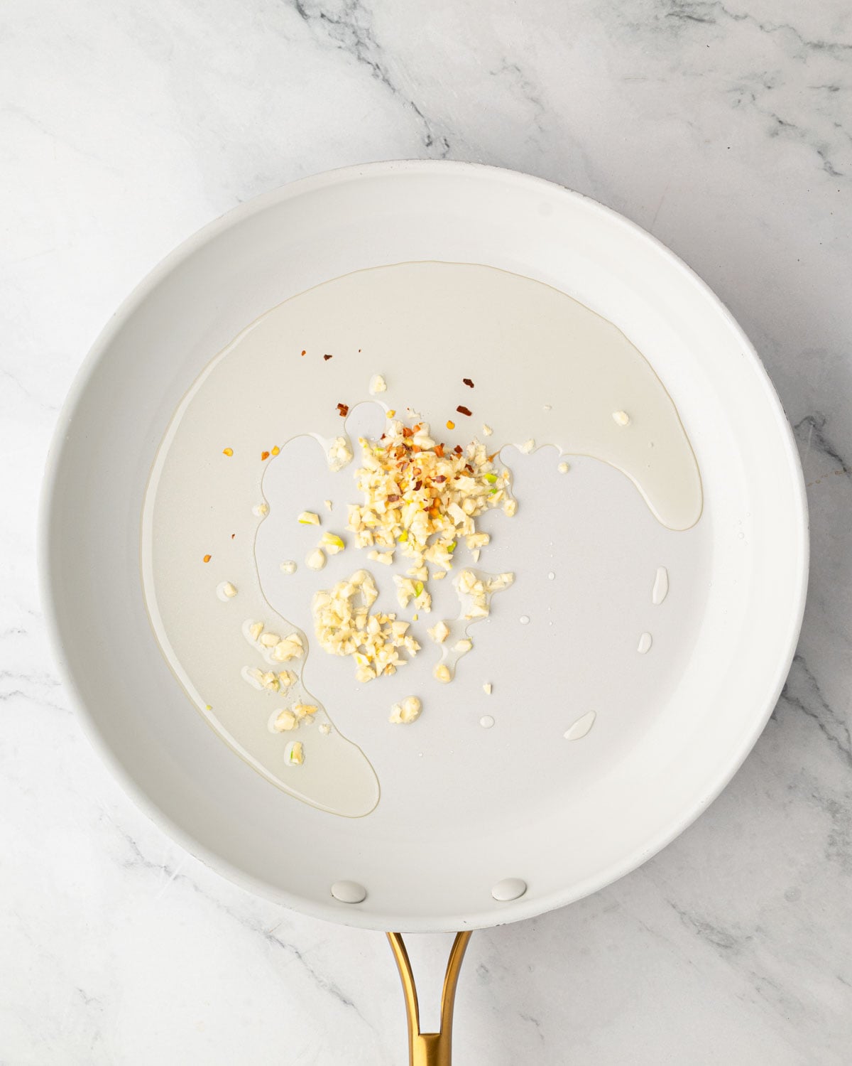 A white skillet with oil, garlic, and chili flakes.