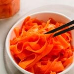 A white bowl of pickled carrots with black chopsticks. A glass jar of more pickled carrots is in the background.