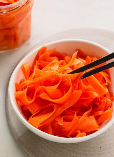 A white bowl of pickled carrots with black chopsticks. A glass jar of more pickled carrots is in the background.