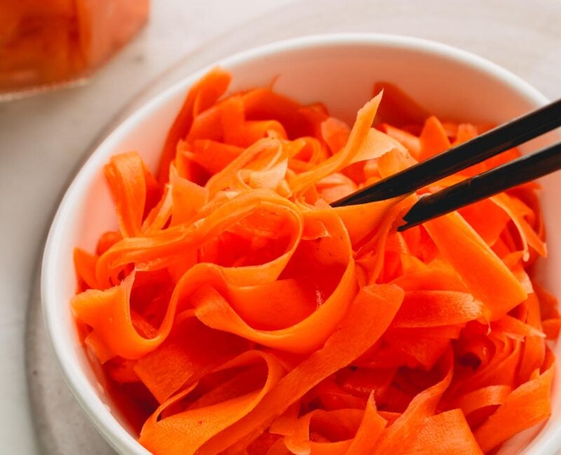 A white bowl of pickled carrots with black chopsticks. A glass jar of more pickled carrots is in the background.