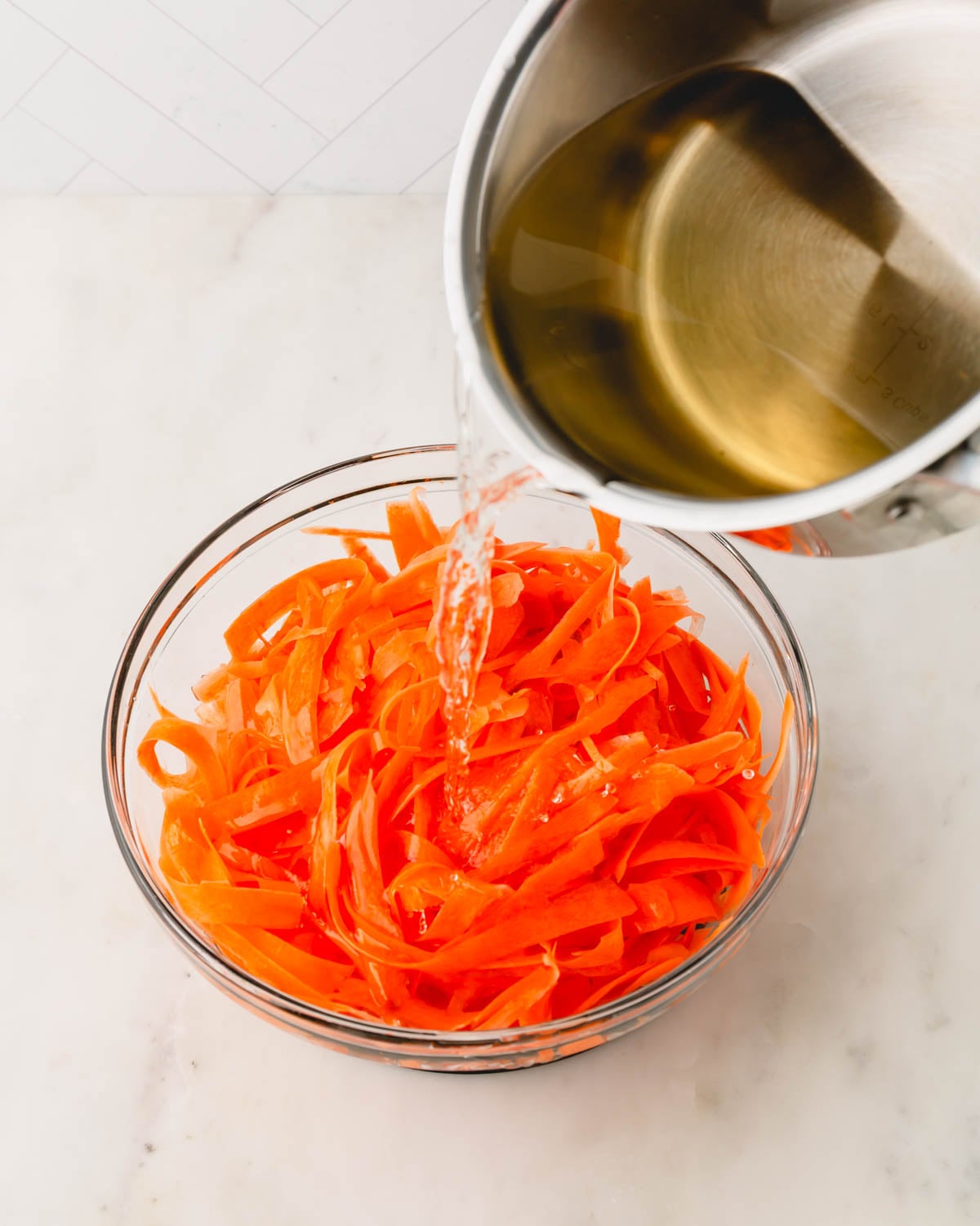 A glass bowl with carrot ribbons and a saucepan of pickling liquid being poured over.
