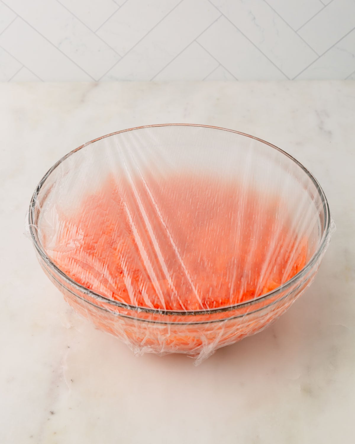 A glass bowl with carrots pickling and plastic wrap over the top.