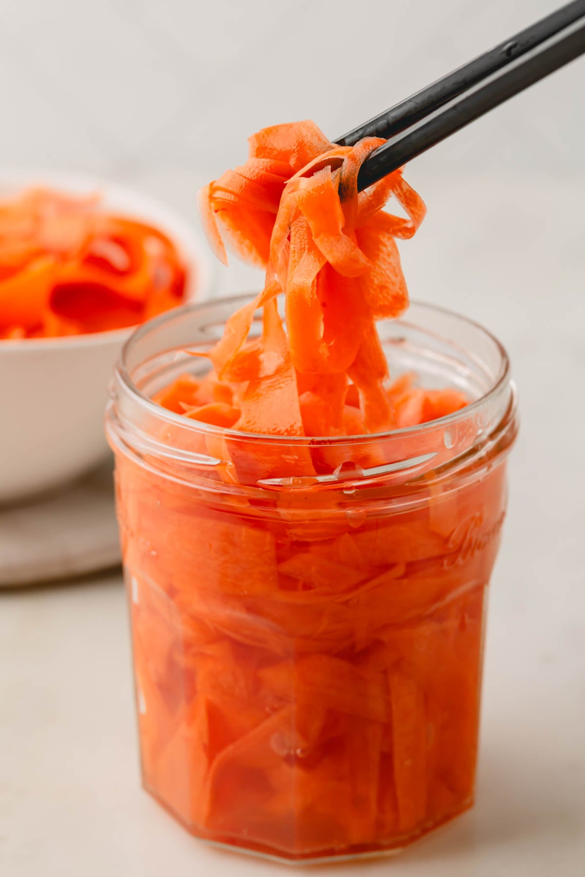 A glass jar of pickled carrots with black chopsticks lifting them up.