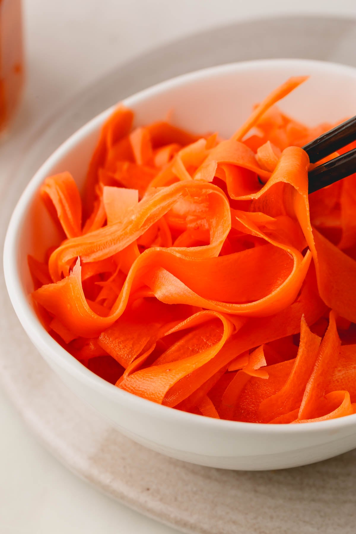 A white bowl of pickled carrot ribbons with black chopsticks picking some up.