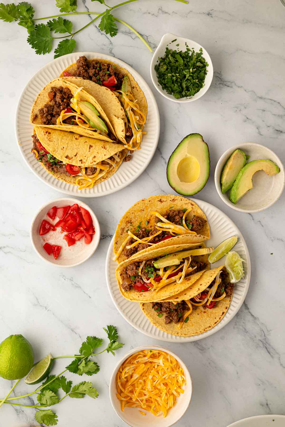 Overhead image of slow cooker ground beef tacos on two white plates with other toppings around it.