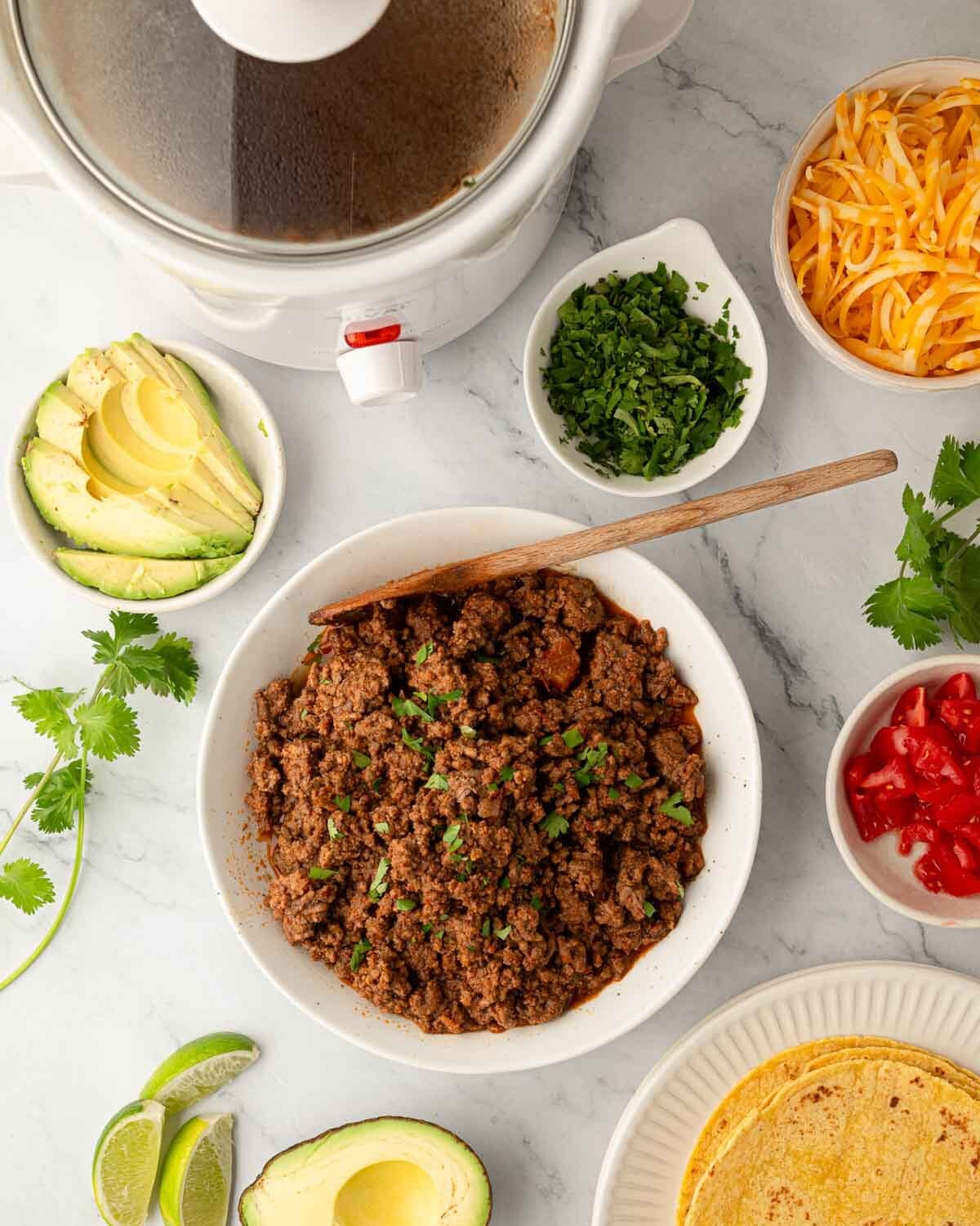 Overhead image of slow cooker ground beef taco meat and toppings for tacos in bowls around it.