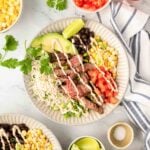Steak bowl in a white plate with small white bowls of other ingredients to the side. A second steak bowl is partially shown in the bottom left corner.