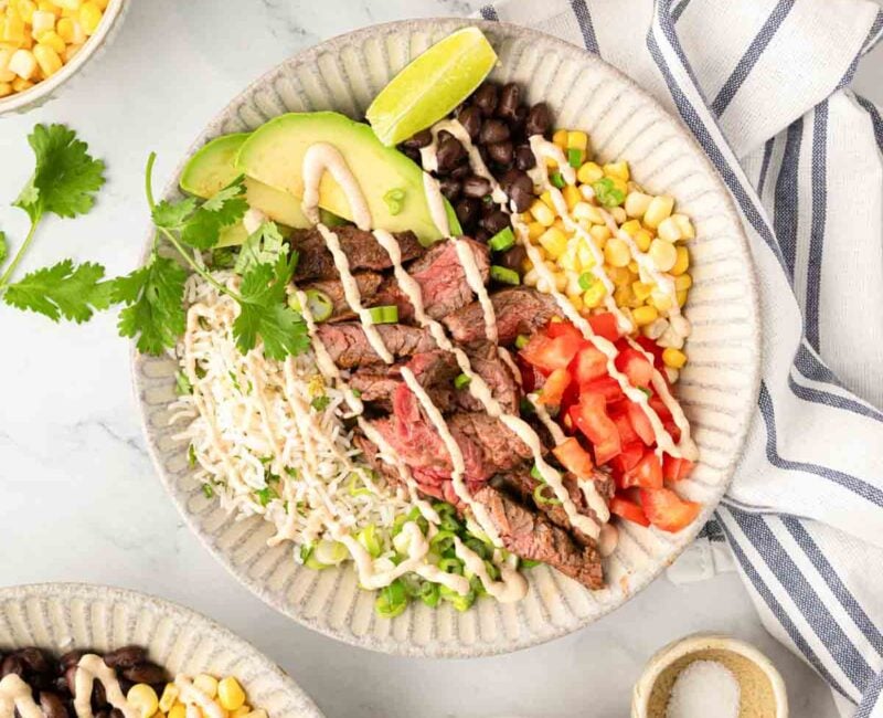 Steak bowl in a white plate with small white bowls of other ingredients to the side. A second steak bowl is partially shown in the bottom left corner.