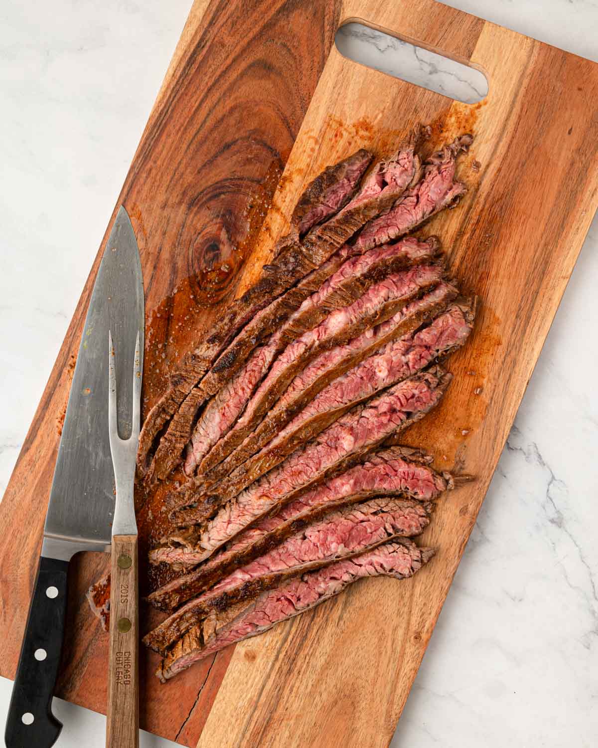 A wooden cutting board with steak sliced against the grain and a knife with a meat fork next to it.