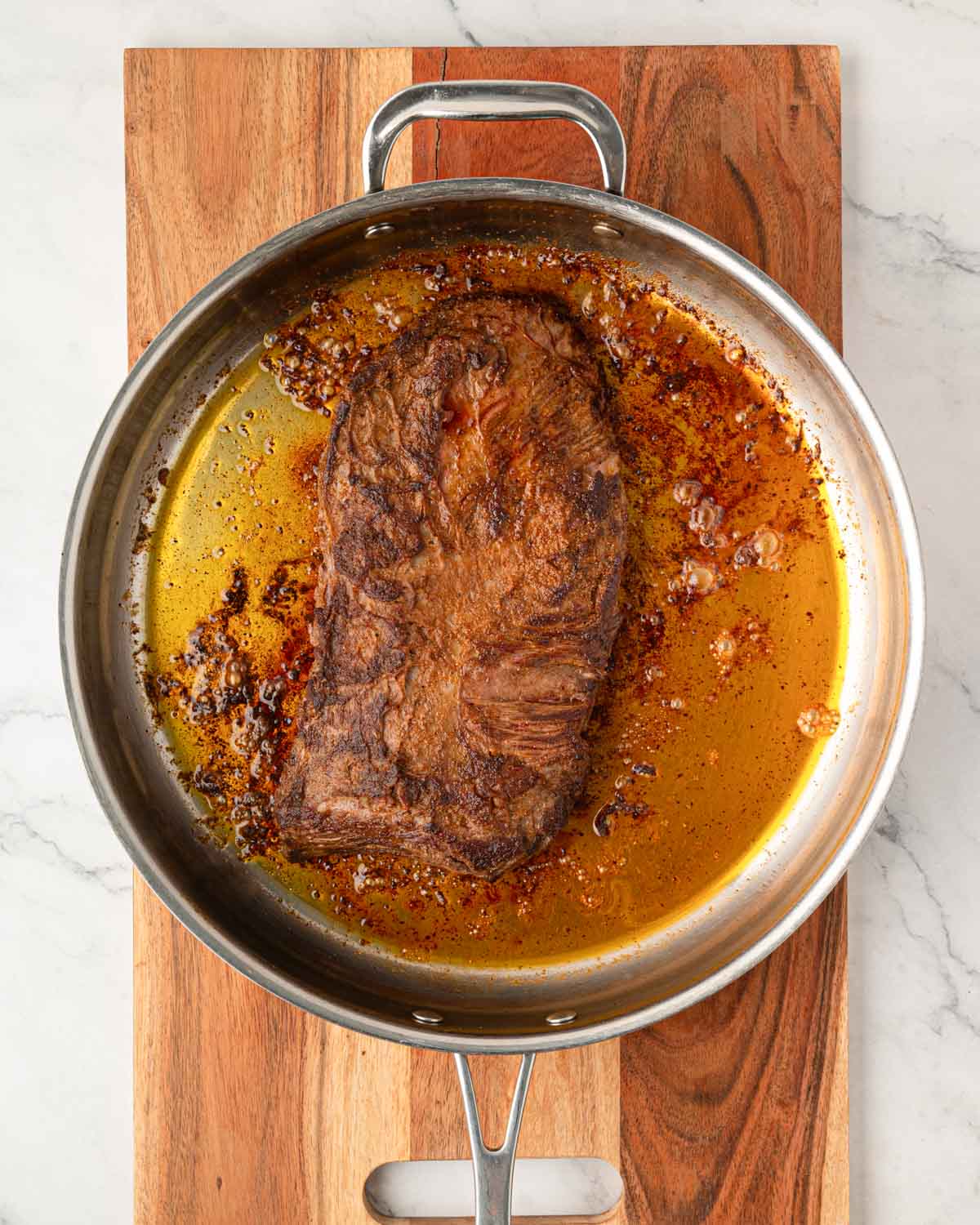 A skillet with steak cooking in oil over a wooden cutting board.